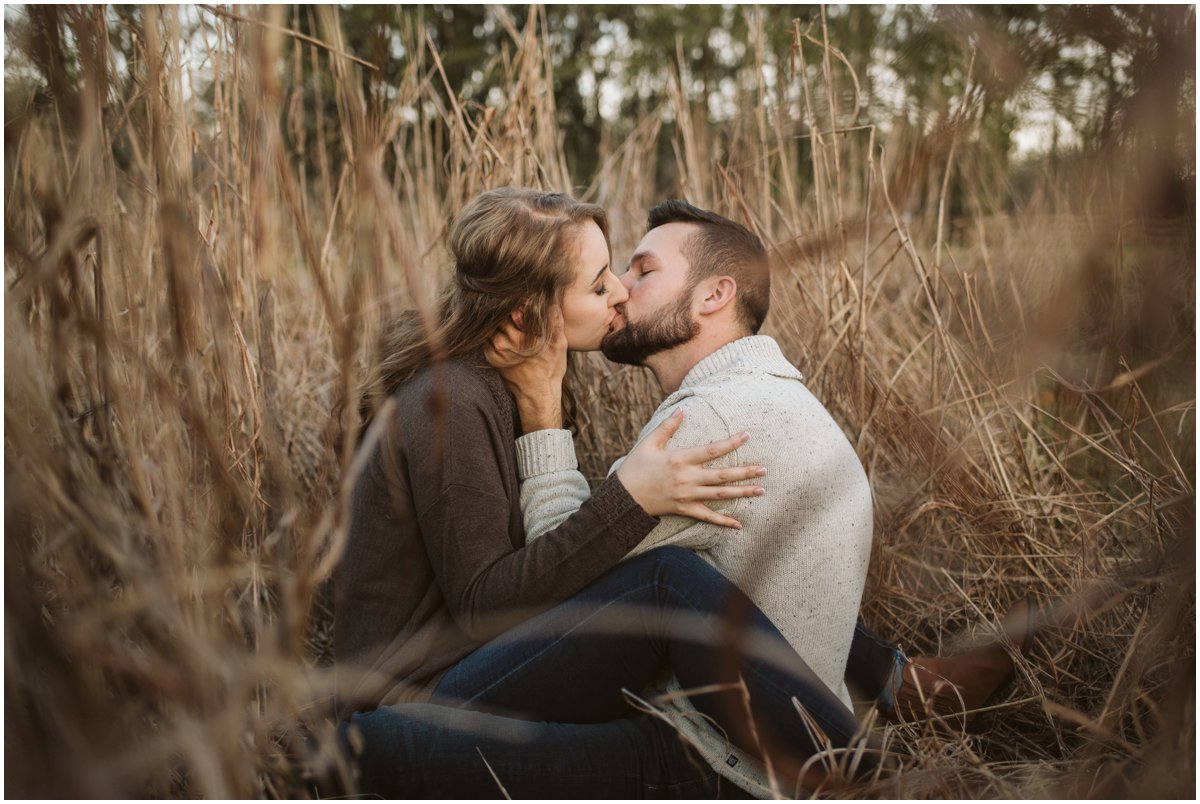 couple sitting in tall grass kissing at belle oaks barn